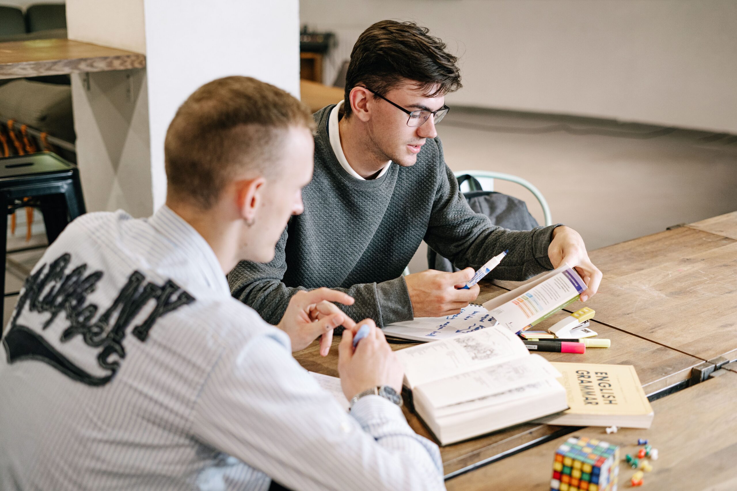 Two young men studying together, focusing on English grammar books in a modern indoor setting.